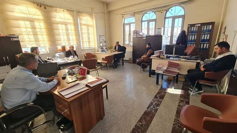 Ofis odasında masalarda oturan beş kişi, dosyalar ve bilgisayarların bulunduğu çalışma ortamında görülüyor.