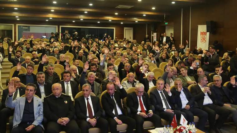 Toplantı salonunda çok sayıda kişi otururken, ön sırada takım elbiseli kişiler ve el kaldıran katılımcılar görülüyor.