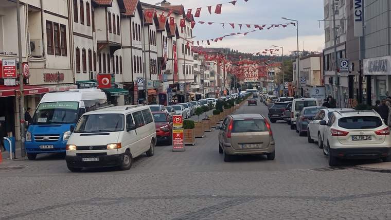 İki yanında park etmiş araçların bulunduğu, dükkânlarla çevrili caddede araçlar ilerliyor; üstte bayrak süslemeleri var.