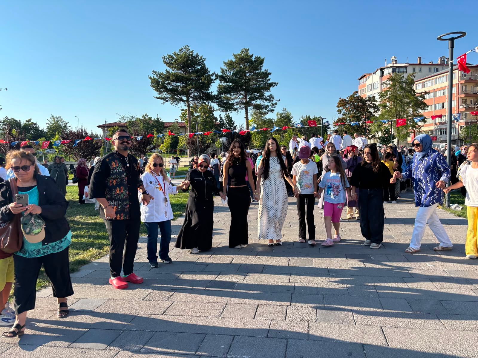 Parkta kırmızı bayrak ve flamalarla süslenmiş alanda yürüyen, sohbet eden kalabalık insanlar görülüyor.