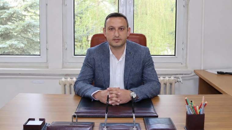 Bir masa başında oturan bir kişi, arkasında büyük pencereler ve yeşil ağaçlar görünmekte.