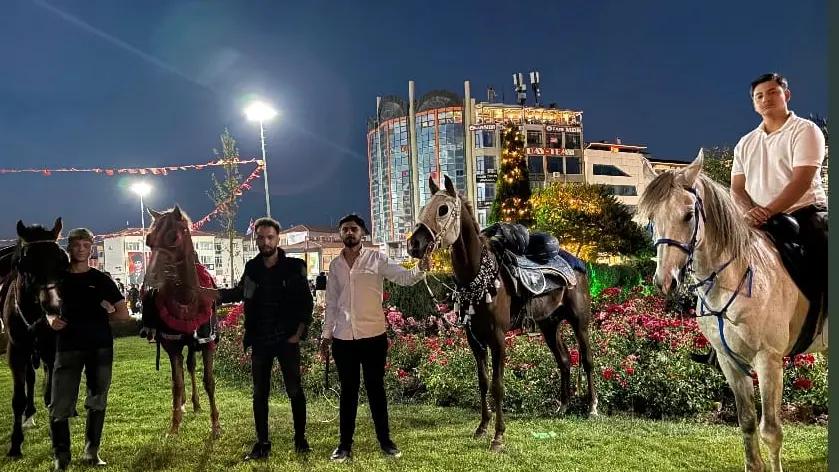 Kent Meydanı'nda at sırtında duran kişiler ve atlar, arka planda binalar ve aydınlatmalar görünüyor.