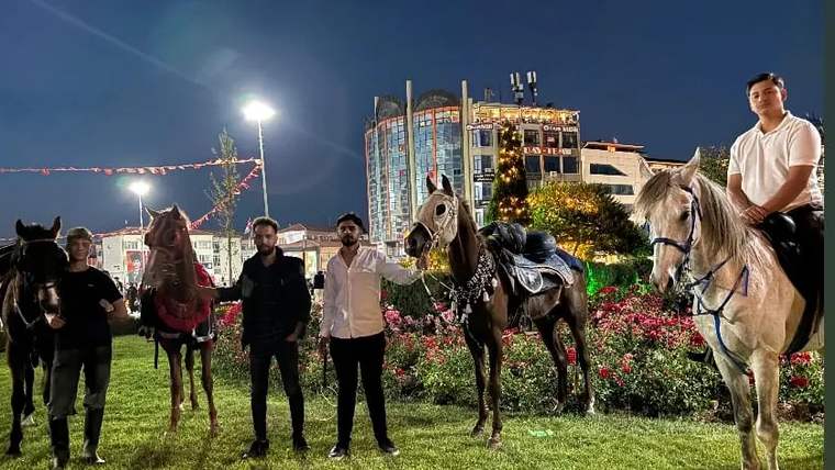 Kent Meydanı'nda at sırtında duran kişiler ve atlar, arka planda binalar ve aydınlatmalar görünüyor.