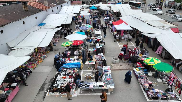 Tente ve şemsiyelerle çevrili açık pazarda, tezgâhlarda giysi ve çeşitli ürünler sergileniyor.