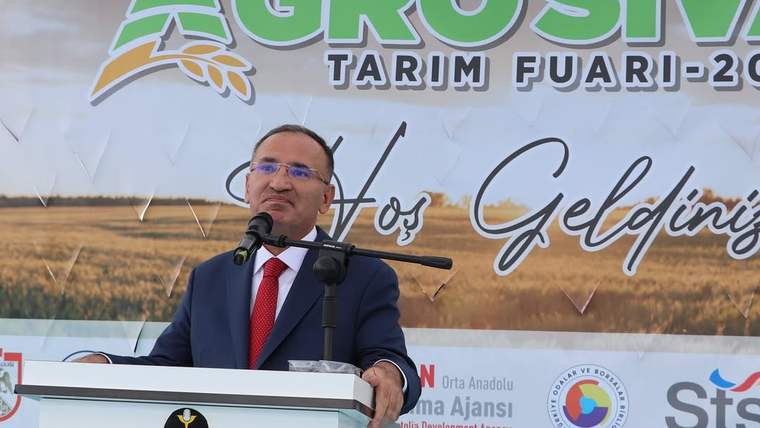 Mikrofonun önünde konuşan bir kişi, arka planda tarım fuarı afişi var.