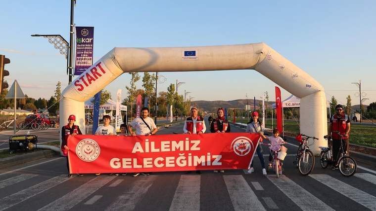 Bisikletliler, üzerinde “AİLEMİZ GELECEĞİMİZ” yazan pankartla başlangıç kemerinin önünde duruyor.