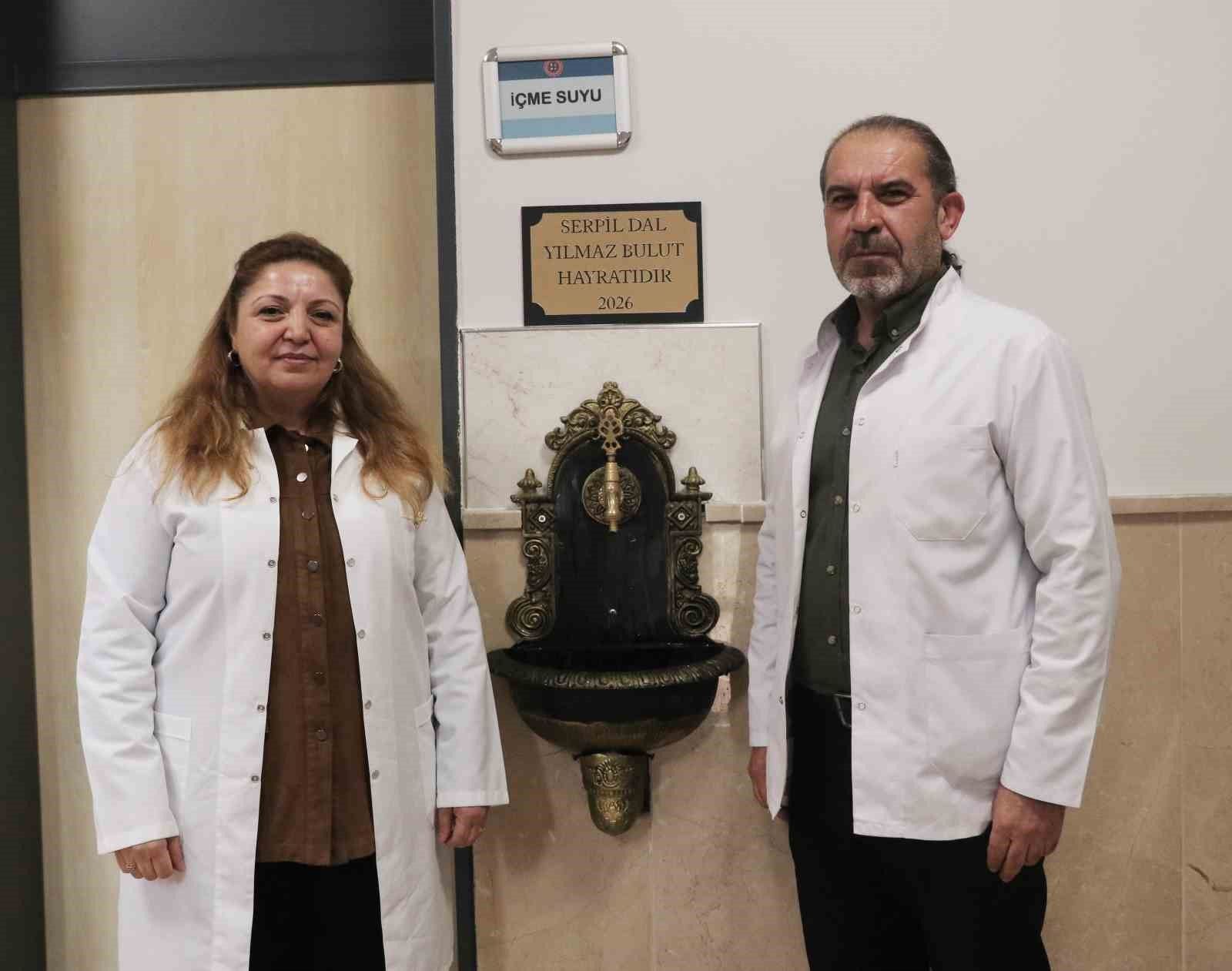 İki beyaz önlüklü kişi, duvara monte süslü çeşmenin yanında duruyor; üstte “İÇME SUYU” yazısı görülüyor.
