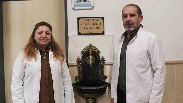 İki beyaz önlüklü kişi, duvara monte süslü çeşmenin yanında duruyor; üstte “İÇME SUYU” yazısı görülüyor.