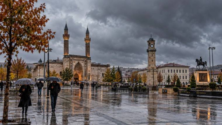 Yağmurlu meydanda şemsiyeli insanlar, iki minareli cami, saat kulesi ve ıslak taş zemin görülüyor.