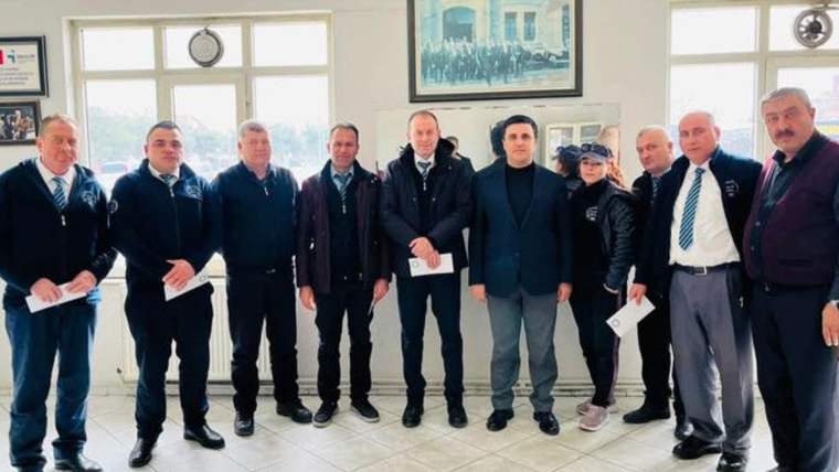 Aydınlık bir salonda, aralarında zarf tutan kişilerin bulunduğu bir grup yan yana duruyor.
