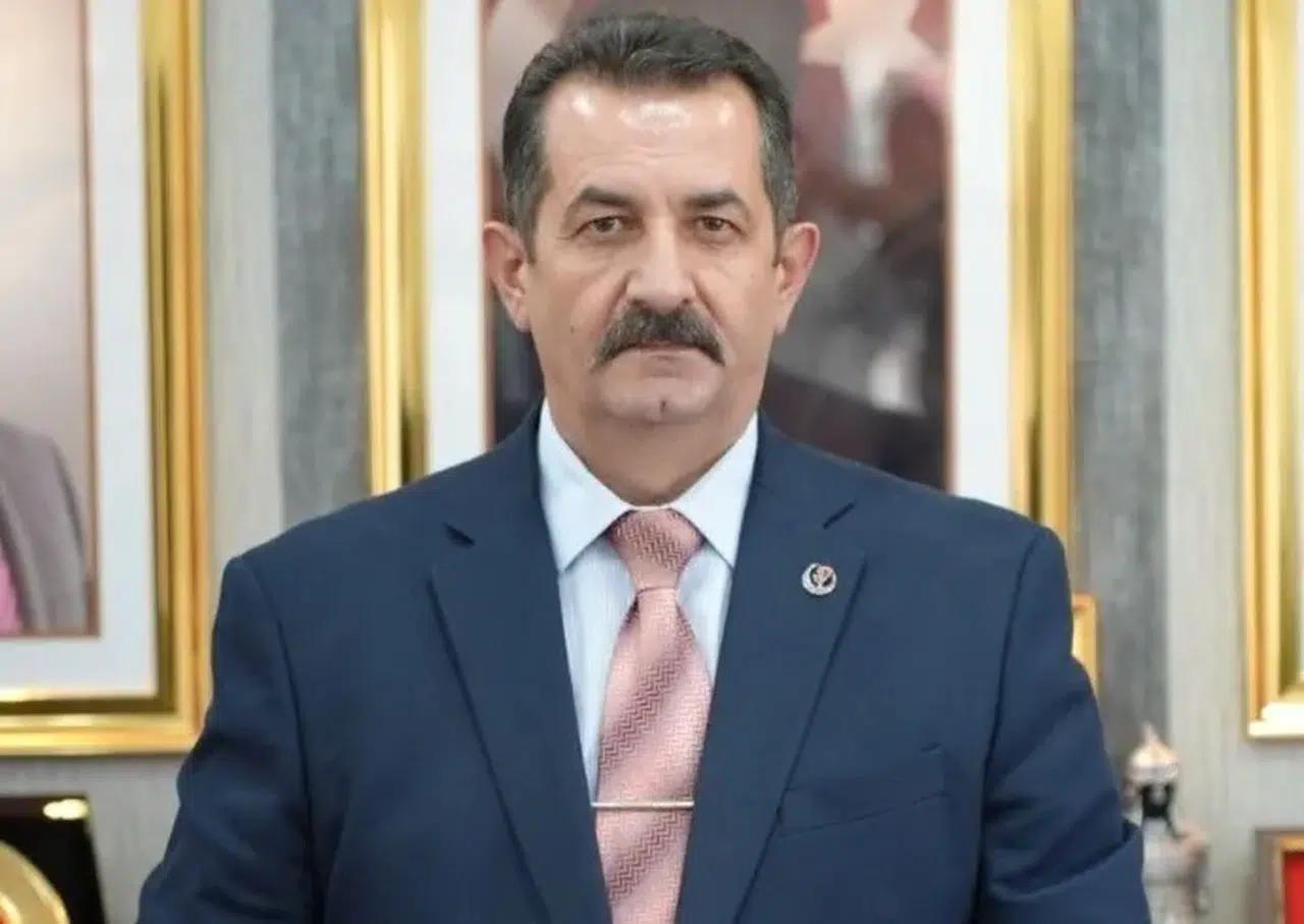 Bıyıklı bir kişi, lacivert takım elbise ve pembe kravatla, çerçeveli tabloların önünde duruyor.