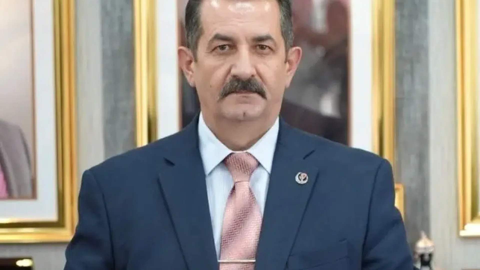 Bıyıklı bir kişi, lacivert takım elbise ve pembe kravatla, çerçeveli tabloların önünde duruyor.