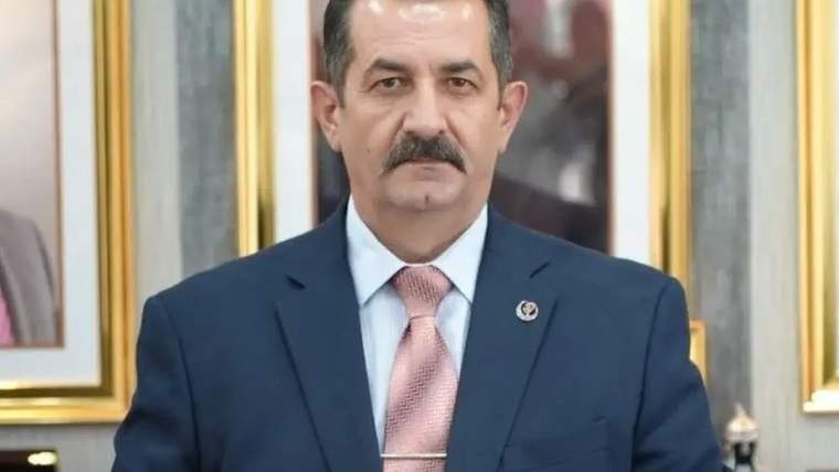 Bıyıklı bir kişi, lacivert takım elbise ve pembe kravatla, çerçeveli tabloların önünde duruyor.