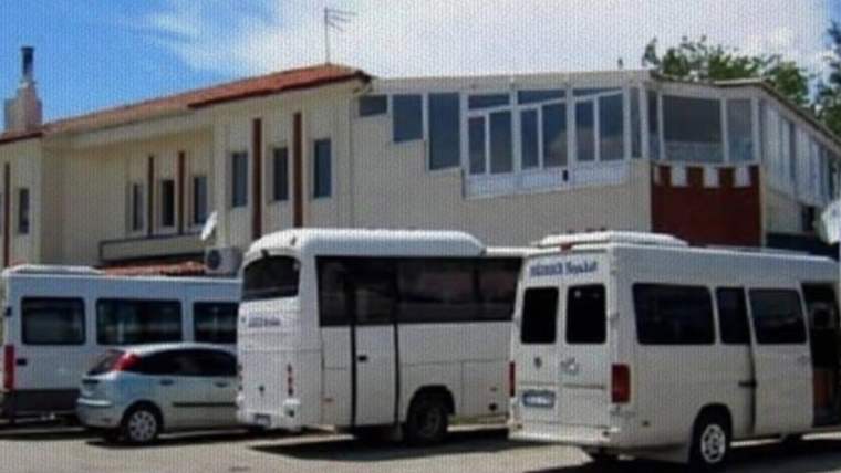 Beyaz minibüs ve otobüsler, önünde park etmiş otomobiller bulunan alçak bir binanın yanında duruyor.