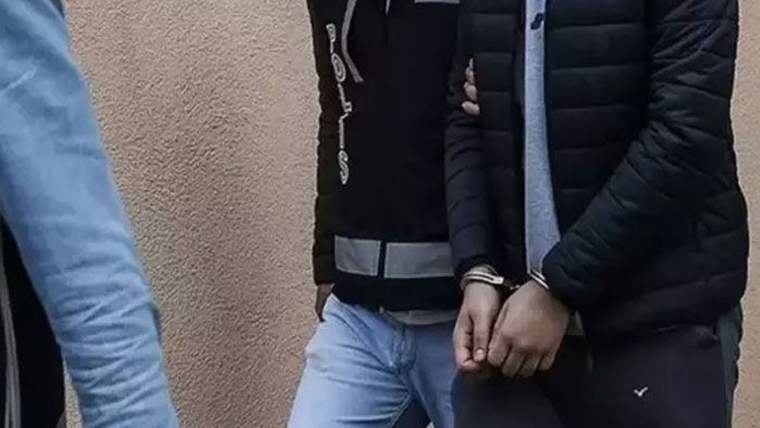 Koyu montlu bir kişi, elleri önde kelepçeli halde duvar önünde duruyor; yanında başka bir kişinin kolu görülüyor.