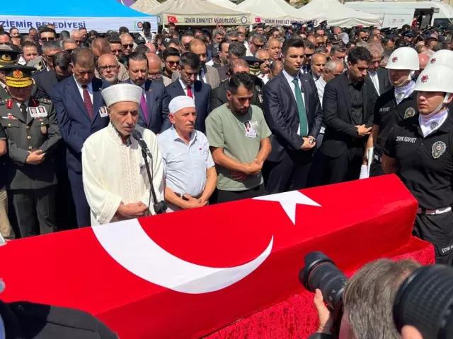Türk bayrağına sarılı tabutun önünde dua eden bir kişi ve etrafında kalabalık bir grup.