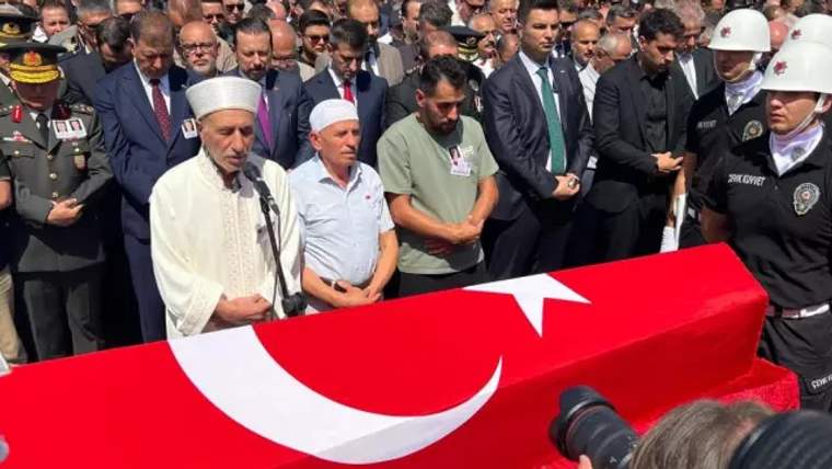 Türk bayrağına sarılı tabutun önünde dua eden bir kişi ve etrafında kalabalık bir grup.