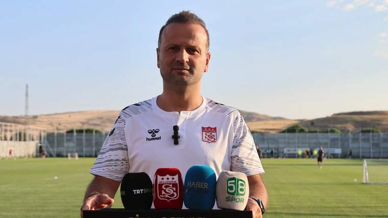 Sivasspor yazılı kürsü önünde, üzerinde takım forması bulunan bir kişi mikrofonlar arasında duruyor.