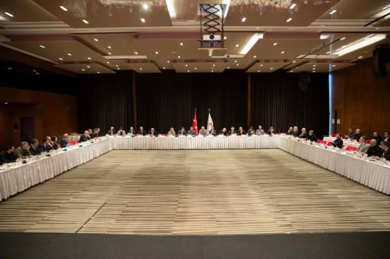 Büyük bir toplantı salonunda, U düzenindeki masalarda oturan kalabalık bir grup görülüyor.