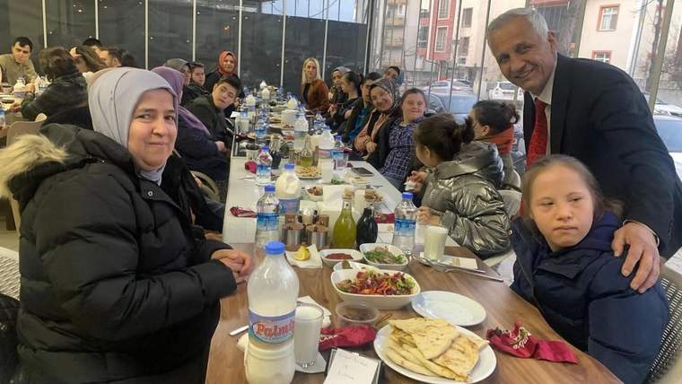 Uzun bir masada oturan kalabalık bir grup, iftar yemeği için hazırlanmış yemeklerin etrafında toplanmış.