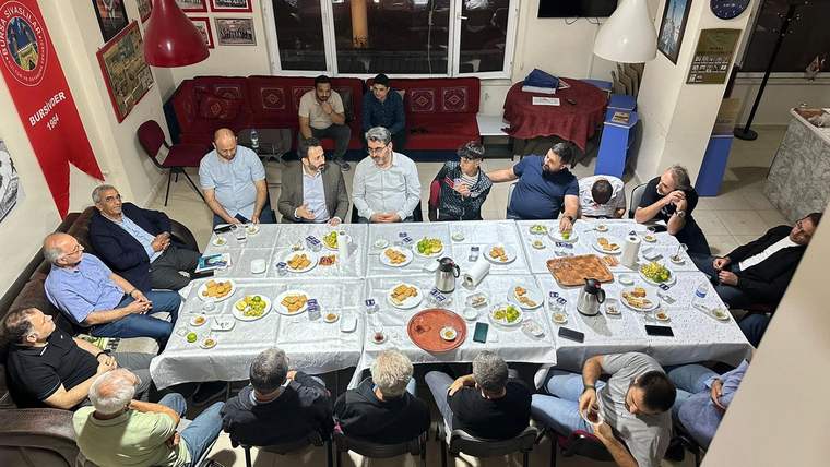 Bir odada uzun masalar çevresinde oturan kişiler, tabaklar, yiyecekler ve içeceklerle birlikte görülüyor.