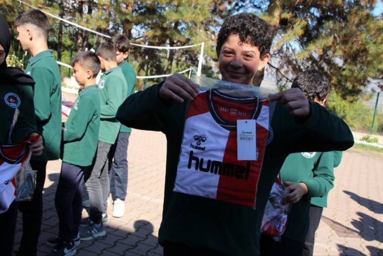 Bir çocuk, üzerinde hummel yazan kırmızı-beyaz futbol formasını tutuyor; çevresinde başka çocuklar bulunuyor.