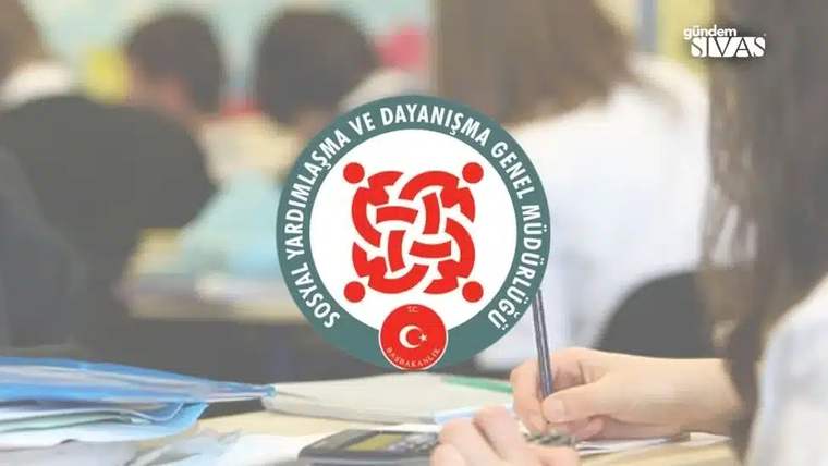 Bir masa üzerinde not defteri ve kalem bulunan bir el, arka planda sınıf ortamı görünmekte.