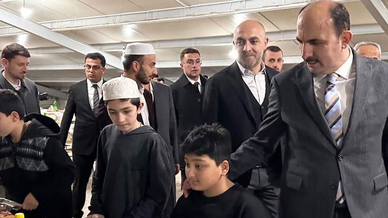 Takım elbiseli yetişkinler, başında beyaz takkeler bulunan iki çocuğun yanında kapalı bir salonda duruyor.
