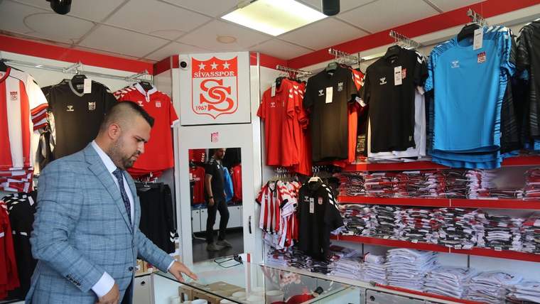 Bir kişi, Sivasspor logolu ürünlerin ve formaların bulunduğu mağazada cam tezgâhın yanında duruyor.