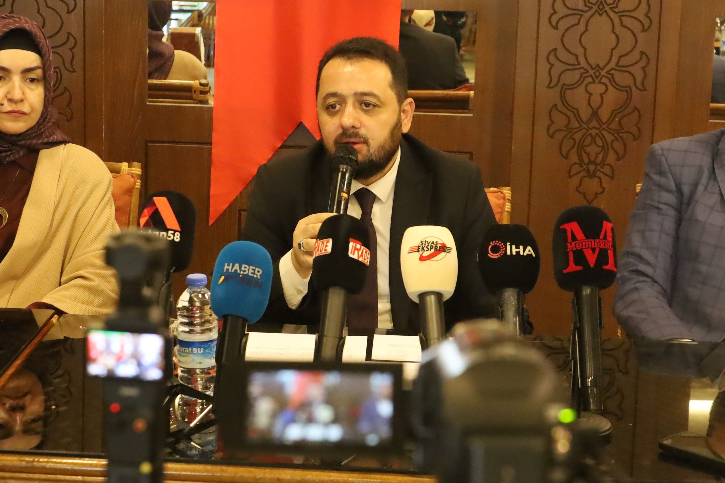 Bir masanın etrafında oturan üç kişi, mikrofonla konuşan bir adamın etrafında toplanmış.