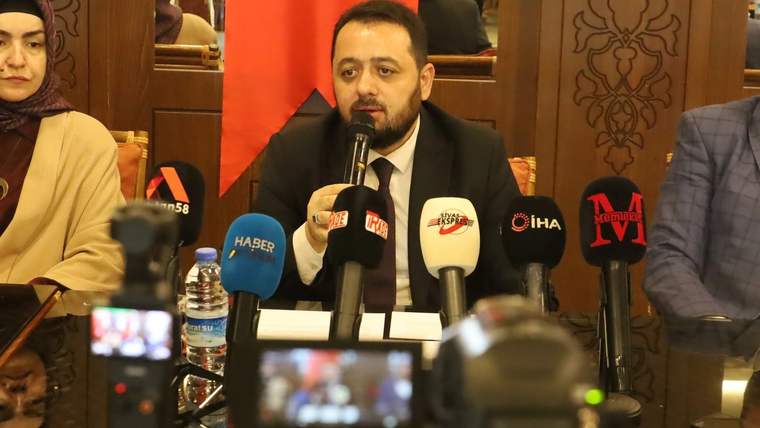 Bir masanın etrafında oturan üç kişi, mikrofonla konuşan bir adamın etrafında toplanmış.