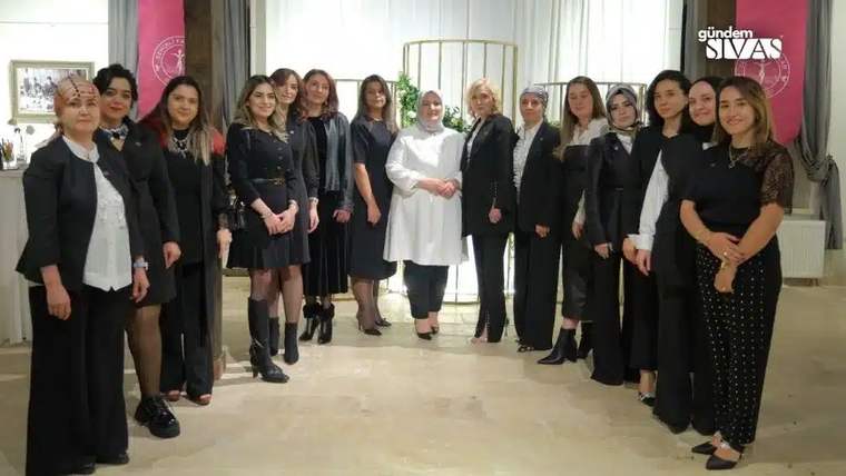 İç mekânda, çoğu siyah giyimli bir grup kadın yan yana duruyor; ortada beyaz kıyafetli bir kadın var.