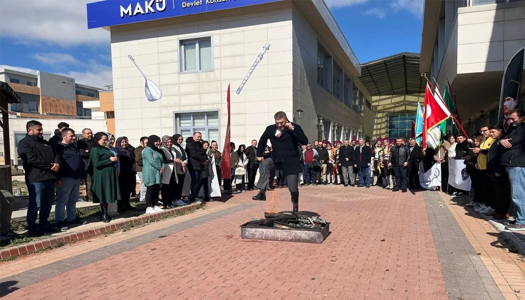 Bir grup insan, Burdur MAKÜ önünde nevruz ateşi etrafında toplanmış, bir kişi demir dövüyor.