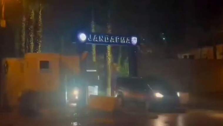 Gece ışıkları altında, üzerinde “Jandarma” yazan girişte araç farları ve ıslak yol görülüyor.