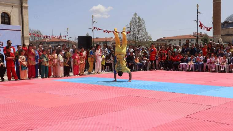 Kırmızı ve mavi minderlerle kaplı alanda bir kişi elleri üzerinde dururken çevresinde kalabalık izliyor.