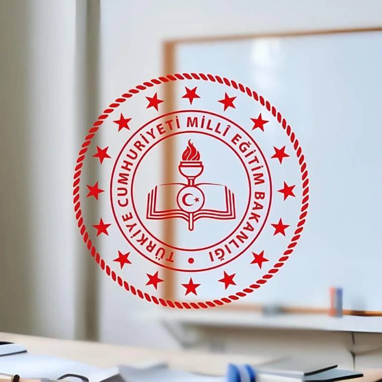 Duvar ve masa üzerinde, Türkiye Cumhuriyeti Millî Eğitim Bakanlığı amblemi bulunan bir sınıf veya ofis ortamı.