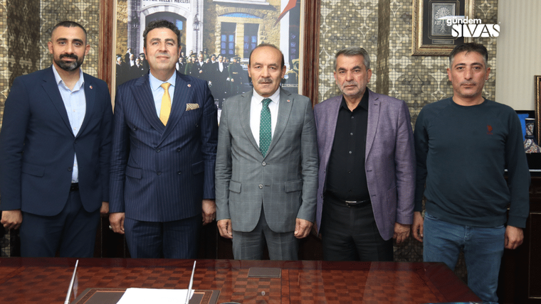Beş kişi, bir masanın önünde poz veriyor; arka planda bir bina ve Türk bayrağı görünmekte.