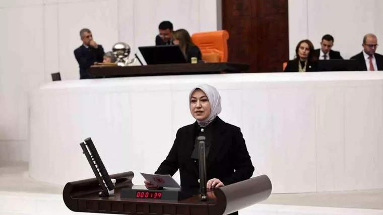 Beyaz başörtülü bir kadın, arkasında oturan kişiler ve elektronik sayaç bulunan kürsüde konuşuyor.