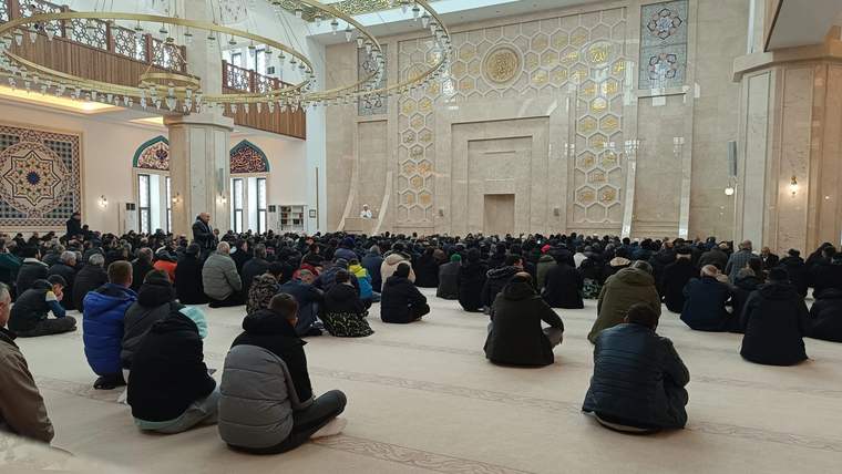 Büyük bir camide, yere oturmuş kalabalık bir grup insan, dua etmekte.