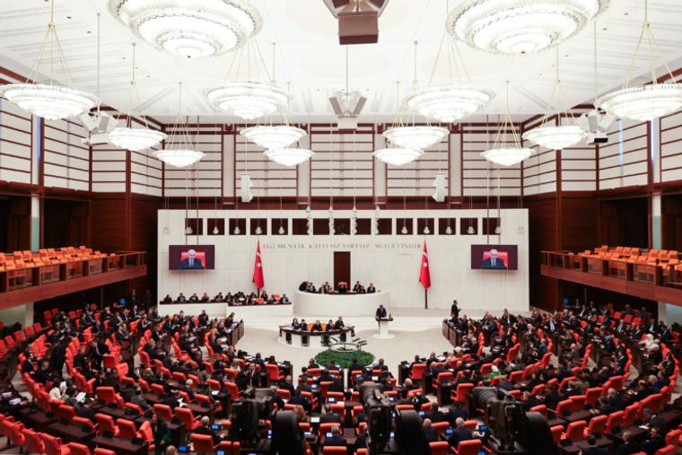 Kırmızı koltuklarla çevrili geniş salonda çok sayıda kişi oturuyor; ön bölümde kürsü ve bayraklar yer alıyor.