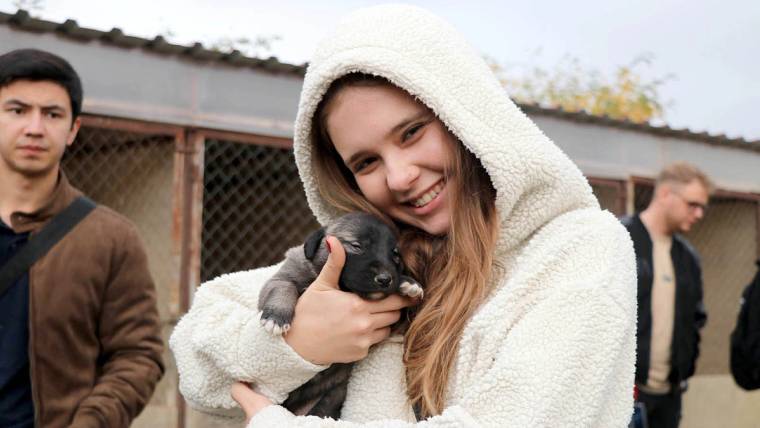 Kapüşonlu bir genç kadın, kucağında siyah bir yavru köpek taşırken çevresinde insanlar görülüyor.