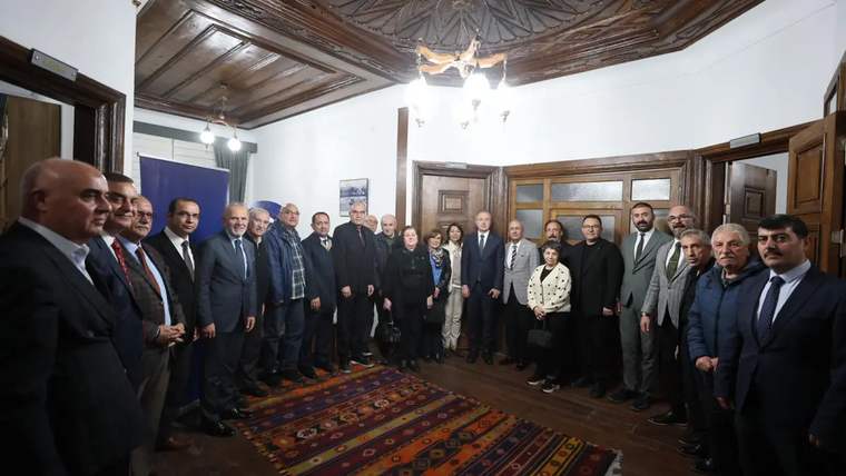 Bir grup insan, bir odada bir araya gelerek poz veriyor. Arka planda ahşap tavan ve halı görünmekte.