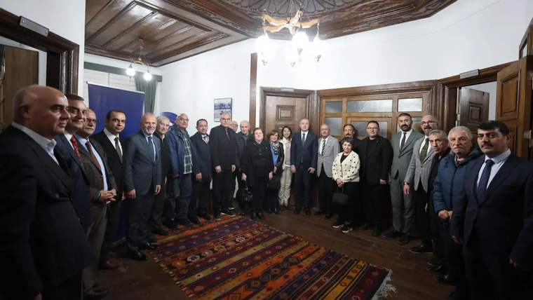 Bir grup insan, bir odada bir araya gelerek poz veriyor. Arka planda ahşap tavan ve halı görünmekte.