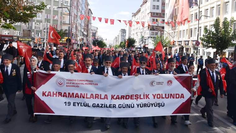 Şehir caddesinde Türk bayrakları taşıyan kalabalık, üzerinde Gaziler Günü yürüyüşü yazılı pankartı taşıyor.