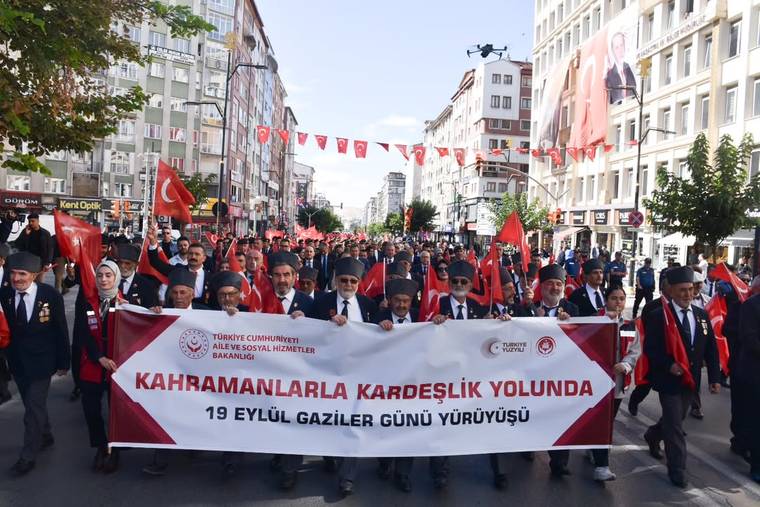 Şehir caddesinde Türk bayrakları taşıyan kalabalık, üzerinde Gaziler Günü yürüyüşü yazılı pankartı taşıyor.