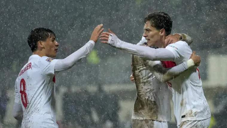 Yağmur altında, beyaz formalar giymiş futbolcular birbirlerine sarılıyor ve sevinç yaşıyor.