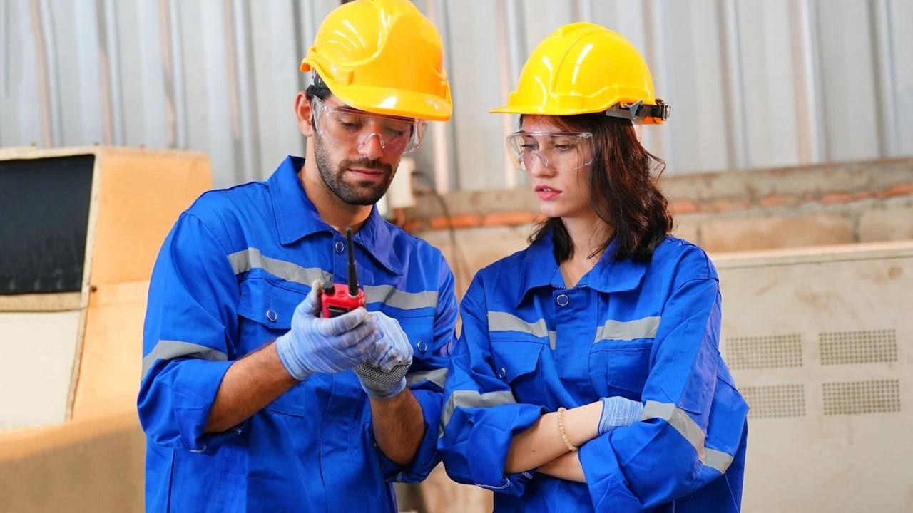 Sarı baret ve mavi iş kıyafetleri giyen iki kişi, eldeki cihazı incelerken görülüyor.