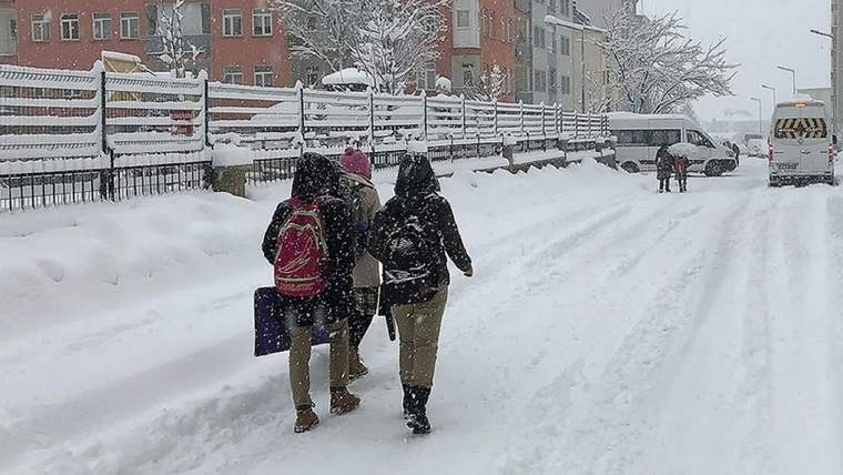 Kar yağışı altındaki yolda sırt çantalı iki kişi yürürken, ileride minibüs ve karla kaplı binalar görülüyor.
