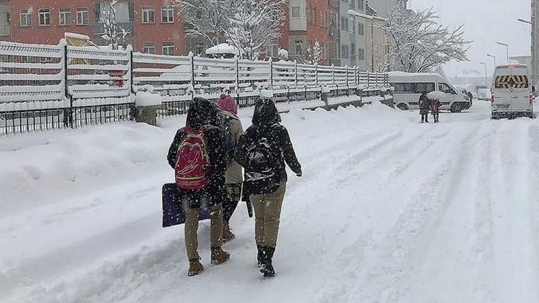 Kar yağışı altında üç kişi, sırt çantaları ve çantalarıyla karlı bir sokakta yürürken görülüyor.