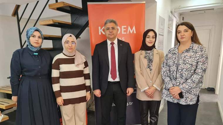 İç mekânda, üzerinde KADEM yazılı turuncu pano önünde dört kadın ve bir erkek yan yana duruyor.